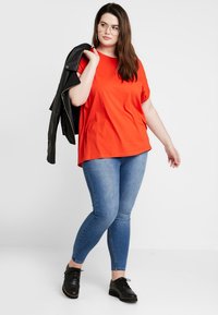 Rød bomull t-skjorte kombinert med lyseblå skinny jeans. Modellen har på seg svarte snørebukser og holder en svart skinnjakke.
