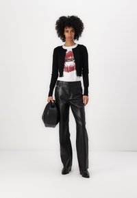 Schwarze gestrickte Strickjacke über einem weißen Grafik-T-Shirt, kombiniert mit hochgeschnittenen schwarzen Lederhosen. Hält eine schwarze strukturierte Handtasche.