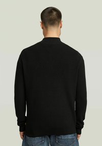 Pull noir en maille côtelée avec col montant, manches longues et coupe décontractée, arborant un discret logo près de l'ourlet.