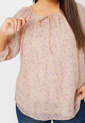 Femme portant un chemisier beige transparent à imprimé floral rose avec un nœud au niveau du col, associé à un jean bleu.
