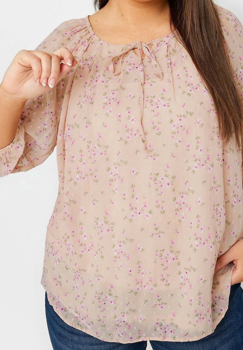 Femme portant un chemisier beige transparent à imprimé floral rose avec un nœud au niveau du col, associé à un jean bleu.