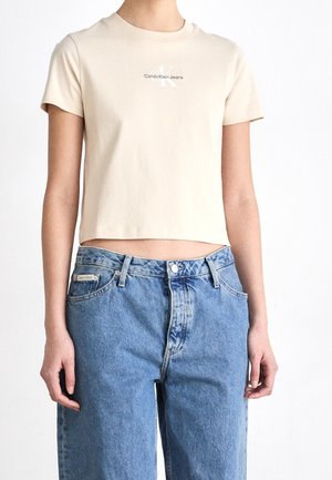 Person iført en kort beige Calvin Klein Jeans t-shirt og højtaljede blå denimjeans mod en ensfarvet hvid baggrund.