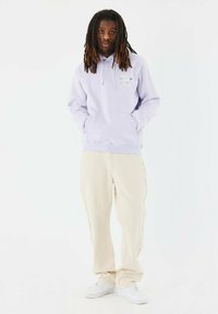 Sudadera lila con bolsillo canguro y detalles gráficos. Pantalones color crema con acentos en las costuras laterales. Calzado: zapatillas deportivas blancas.