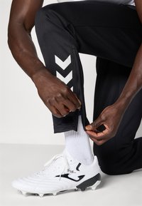 Kezek cipzárt igazítanak fekete sportnadrágon, fehér focicipő és fehér zokni felett, egy térdelő személyen.