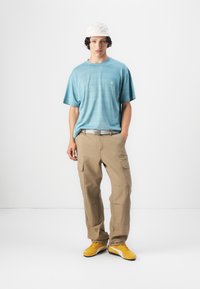 Lyseblå stripete t-skjorte, beige cargo-bukser med sidelommer, grå belte og gule joggesko. Modellen står foran en ensfarget hvit bakgrunn.