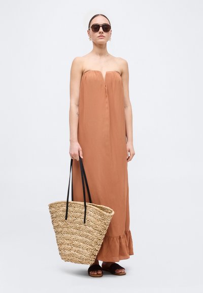 Kvinde iført stropløs brun maxikjole og mørke solbriller, holder en stor flettet stråtaske med sorte håndtag, stående i sandaler.