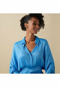 Ljusblå blus i mjukt tyg med knäppning vid kragen och lösa, flödande ärmar. Positionerad mot en neutral bakgrund.