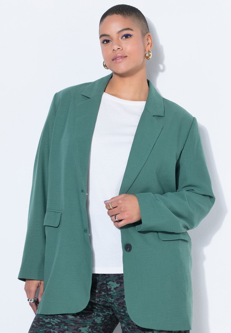 Person mit kurzen Haaren, die goldene Creolen, einen grünen, übergroßen Blazer, ein weißes Hemd und camouflagemusterte Hosen trägt und vor weißem Hintergrund posiert.