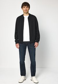 Svart bomberjacka med rund hals, framdraget blixtlås, bärs över en vit skjorta, kombinerad med mörkblå jeans och vita sneakers, stående mot en enkel bakgrund.