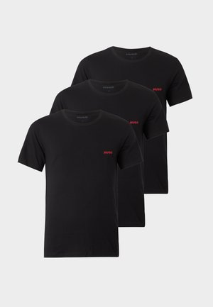 Homme debout pieds nus portant un t-shirt noir et un pantalon de jogging avec petits logos rouges Hugo, les mains dans les poches, sur un fond blanc uni.