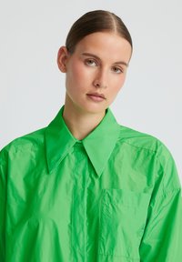 Chemise surdimensionnée verte en tissu léger et brillant. Dotée d'un grand col et d'une poche avant sur le côté gauche.