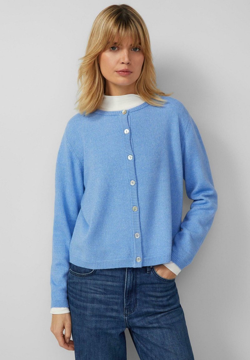 Frau trägt eine hellblaue, geknöpfte Strickjacke über einem weißen Oberteil, kombiniert mit dunkelblauen Jeans, stehend vor einem schlichten hellgrauen Hintergrund.