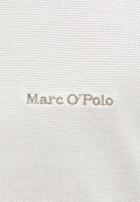 Μπεζ πλεκτό ύφασμα με κεντημένο το 'Marc O'Polo' σε ανοιχτό καφέ κλωστή, στο κέντρο του υφάσματος.