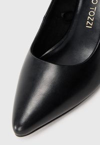 Chaussure femme en cuir noir à bout pointu, avec un devant bas et une texture lisse, montrant partiellement le texte de la semelle intérieure de marque.
