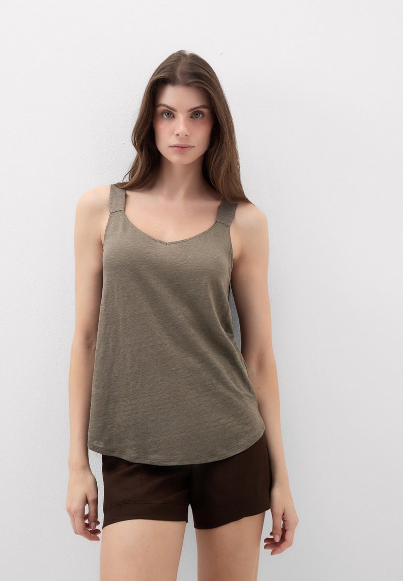 Jonge vrouw met lang bruin haar, die een mouwloze olijfgroene top en donkerbruine korte broek draagt, staand tegen een effen witte muur.