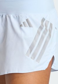 Svetlo modra atletična krilo s gladko teksturo, stranskimi razporki in odsevnim sivim logotipom s tremi diagonalnimi črtami na spodnjem robu.