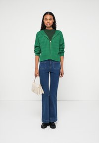 Zöld kötött, cipzáras kardigán, texturált mintával, flared farmernadrággal párosítva. A modell egy krémszínű kézitáskát tart. Fekete cipők egészítik ki a szettet.