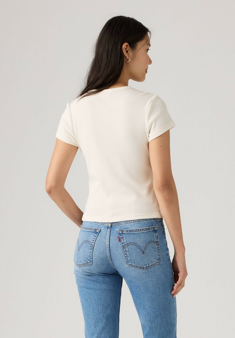 Femme aux longs cheveux foncés portant un t-shirt blanc à manches courtes côtelé et un jean bleu taille haute, debout de dos.