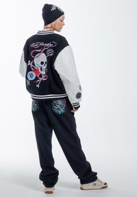 Black and white varsity jacket with embroidered skull and colorful designs on back; black pants with dragon patterns; beige sneakers.