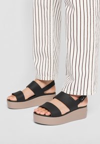 Schwarze Plateausandalen mit zwei breiten Riemen und einer beigen Sohle. Glattes Material mit einem verstellbaren Fersenriemen, der kleine Hardware-Details aufweist.