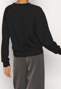 Personne portant un pull noir à manches longues et un pantalon gris ample, vue de dos sur un fond blanc.