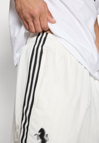 Pantalones cortos deportivos blancos con rayas negras a los lados, con un logo bordado y una tela texturizada. Detalle de cintura elástica visible.