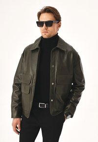 Tumed roheline nahkjope, millel on nööbid ees, kaks taskut ning kraega kaelus, kantud musta kõrge kaelusega särkide kohal.