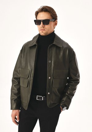 Chaqueta de cuero verde oscuro con cierre de botones en la parte delantera, dos bolsillos de parche y cuello, llevada sobre un suéter de cuello alto negro.