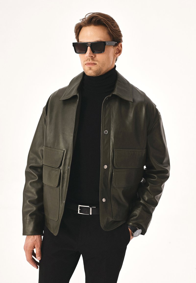 Tumed roheline nahkjope, millel on nööbid ees, kaks taskut ning kraega kaelus, kantud musta kõrge kaelusega särkide kohal.