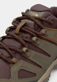 Zapato de senderismo marrón oscuro con parte superior texturizada, detalles en naranja y cordones marrones. Cuenta con puntera reforzada y diseño de suela flexible para tracción.