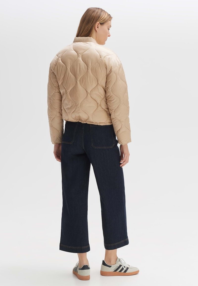 Beige gesteppte Jacke mit rundem Kragen, kombiniert mit dunkelblauen weit geschnittenen Jeans und grauen Sneakers mit schwarzen Streifen und Gummisohlen.
