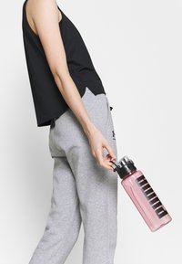 En person i en svart ärmlös topp och grå sweatpants håller en rosa Puma-vattenflaska i sin ögla mot en ljus bakgrund.