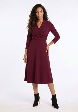 Femme aux longs cheveux bouclés portant une robe midi portefeuille bordeaux et des bottines noires à talons, debout devant un fond blanc.