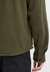Chaqueta de forro polar verde oliva con una superficie texturizada, puños abotonados y un dobladillo ligeramente curvado. Combinada con pantalones oscuros.