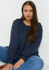 Blusa a maniche lunghe color blu navy con colletto arricciato, tessuto testurizzato, rifinitura in pizzo decorativo e vestibilità comoda, abbinata a jeans in denim.
