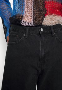 Black denim jeans with a smooth texture, featuring front pockets, belt loops, and a metal button closure. Worn with a colorful, knitted top.