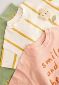 Tres camisetas de algodón: una color crema con rayas amarillas y detalle de flores, una rosa con el texto "smile" y una verde como fondo.