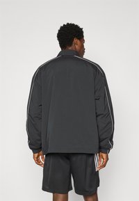 Veste noire à coupe décontractée, avec des rayures blanches sur les côtés et des poignets élastiques. Coutures visibles et col rabattu. Portée avec un short noir.