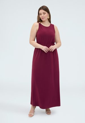 Femme portant une robe longue bordeaux sans manches et des talons beiges, debout les mains jointes et les yeux fermés sur un fond clair uni.