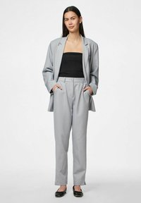 Gaiši pelēks piegriezējuzvalks ar plašiem atlokiem, divām priekšējām kabatām un augsto jostasvietu biksēm. Melna pieguļoša blūze un plakani melni apavi papildina tēlu.