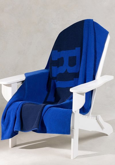 Μπλε φλις ρίχτης κουβέρτα ριγμένη πάνω σε μια λευκή ξύλινη καρέκλα. Διαθέτει ένα μείγμα σκούρου μπλε και ανοιχτού μπλε τμημάτων με μεγάλα γράμματα.
