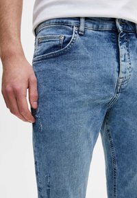 Hombre vistiendo jeans de mezclilla azul claro con sutiles detalles desgastados y una camisa blanca, con la mano relajada a un lado contra un fondo blanco.