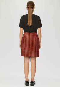 Zwart T-shirt met korte mouwen, gecombineerd met een rok in rood en zwart houndstooth-patroon. Model draagt zwarte schoenen; glad haar is naar achteren gestyled.