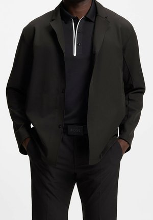 Mann trägt schwarze offene Jacke mit Knöpfen über schwarzem Polo mit weißem Reißverschlussdetail, schwarze Hose und schwarzer Gürtel mit "BOSS" Logo.