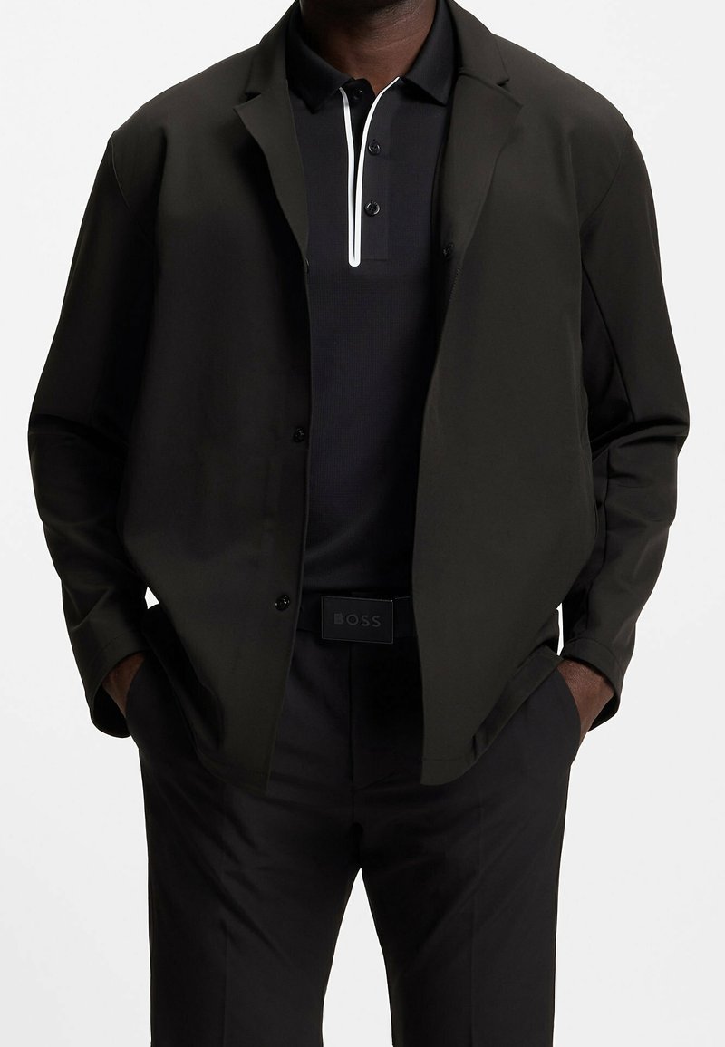 Homme portant une veste noire ouverte à boutons sur un polo noir avec un détail de zip blanc, pantalon noir et ceinture noire avec le logo "BOSS".