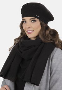Béret noir et écharpe noire en tissu doux, drapés de manière décontractée. Le mannequin porte un manteau gris, mettant en avant une texture lisse et un design élégant.