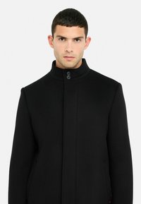 Cappotto di lana nero con colletto alto, caratterizzato da una silhouette dritta, due bottoni al collo e tasche laterali. Design semplice e moderno.