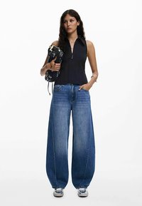 Must sleeveless top kraega, kombineeritud laia lõikega siniste teksapükstega. Model hoiab mustrilist kotti ja kannab hõbedaseid tossusid.