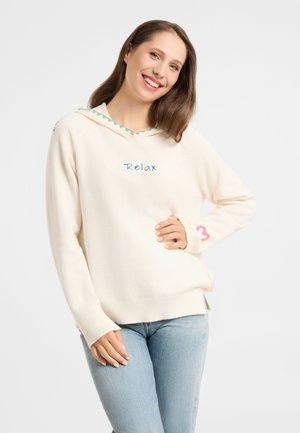 Smiling woman with long brown hair wears cream hoodie with "Relax" text and light blue jeans, posing with one hand on hip and white background.