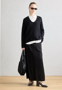 Maglione nero in pelliccia, camicia bianca con colletto sotto, gonna maxi nera e borsa a tracolla plissettata nera. Scarpe nere opache con cinturini alla caviglia.